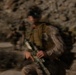 A motion-blurred photograph depicts multiple armed military personnel moving through a rocky, arid terrain. The central figure is most prominent, carrying a rifle and wearing tactical gear including a vest and what appears to be eye protection or a mounted device on the head. At least two additional soldiers are visible in the background, suggesting a patrol or movement operation.