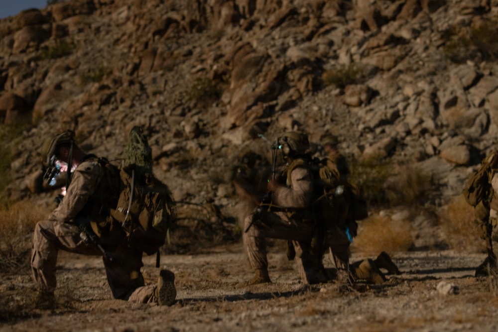 The image shows multiple military personnel in desert camouflage uniforms kneeling on arid, rocky terrain at the base of a rocky hillside. The soldiers appear to be in a tactical formation, carrying full combat gear including backpacks and weapons. The setting suggests a desert training exercise or field operation.