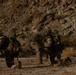 The image shows multiple military personnel in desert camouflage uniforms kneeling on arid, rocky terrain at the base of a rocky hillside. The soldiers appear to be in a tactical formation, carrying full combat gear including backpacks and weapons. The setting suggests a desert training exercise or field operation.