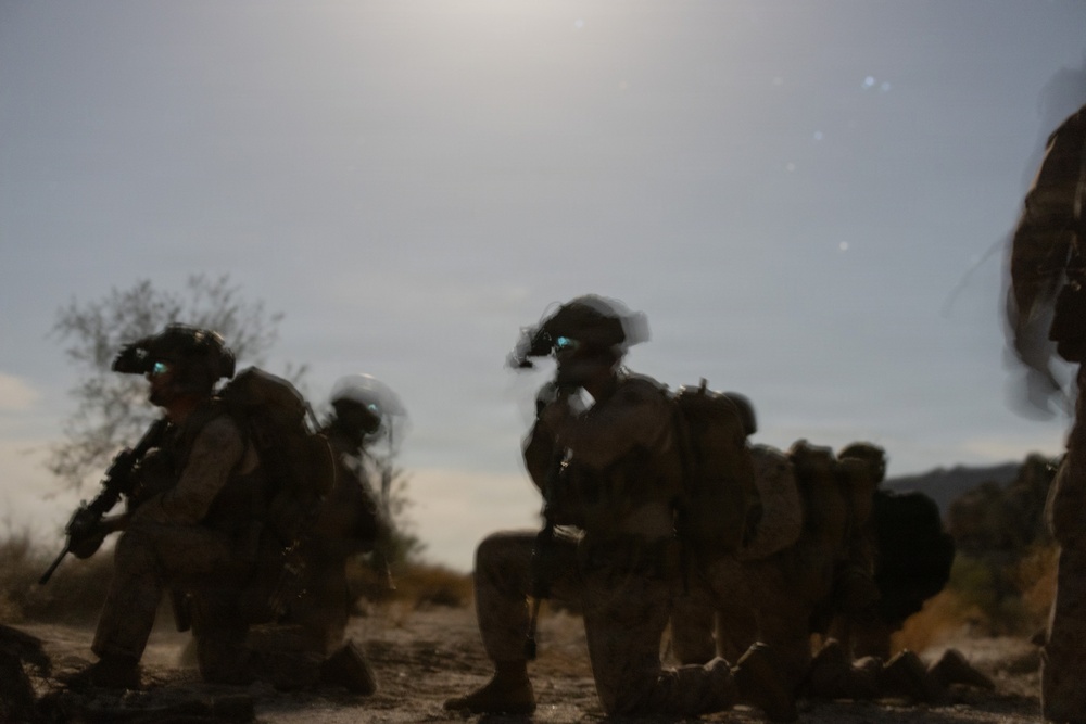 The image shows a group of heavily equipped military personnel in a desert or arid environment, appearing to be in a tactical formation or patrol. The soldiers are wearing full combat gear including helmets, body armor, backpacks, and tinted protective eyewear. The image is motion-blurred and backlit by a bright sun, giving it a hazy, low-contrast appearance.