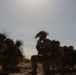 The image shows a group of heavily equipped military personnel in a desert or arid environment, appearing to be in a tactical formation or patrol. The soldiers are wearing full combat gear including helmets, body armor, backpacks, and tinted protective eyewear. The image is motion-blurred and backlit by a bright sun, giving it a hazy, low-contrast appearance.