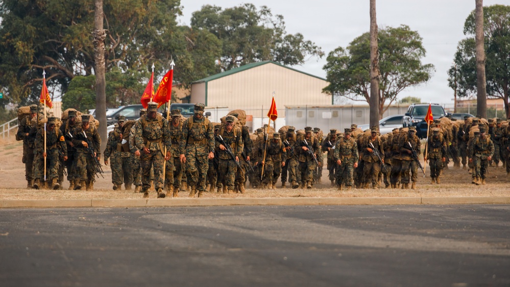A large formation of U.S. Marines in woodland MARPAT camouflage uniforms conducts a combat march or hike carrying full gear, rucksacks, and rifles. Multiple red unit guidon flags are visible among the formation, indicating organized unit movement. The setting appears to be a military installation, likely on the West Coast based on the vegetation and terrain.