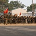 A large formation of U.S. Marines in woodland MARPAT camouflage uniforms conducts a combat march or hike carrying full gear, rucksacks, and rifles. Multiple red unit guidon flags are visible among the formation, indicating organized unit movement. The setting appears to be a military installation, likely on the West Coast based on the vegetation and terrain.