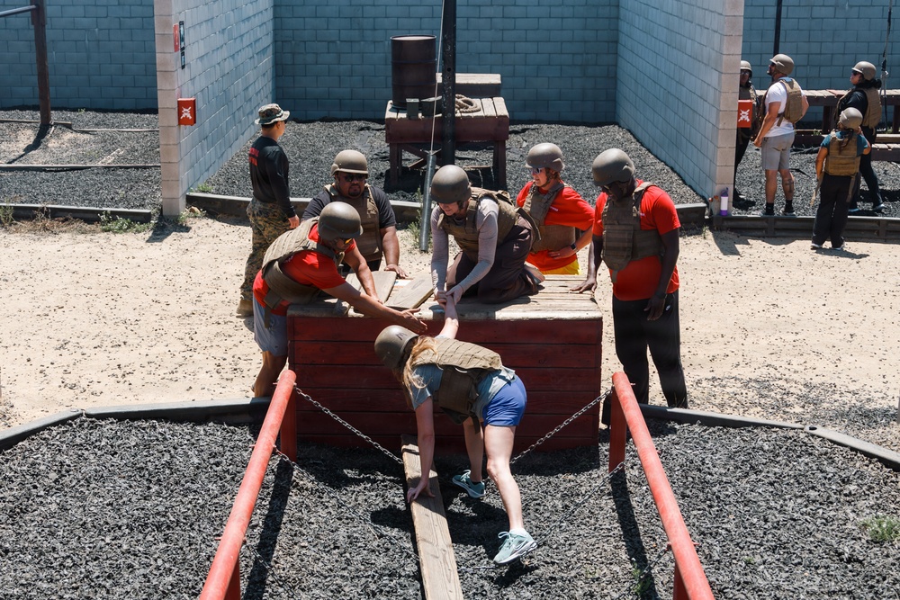 A group of civilians wearing helmets and tactical vests are participating in a military-style team-building or obstacle course exercise. Several participants in red shirts are helping pull a person up over a wooden platform obstacle surrounded by a gravel pit with red rails and chains. Additional participants and observers are visible in the background near a cinder block wall.
