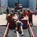 A group of civilians wearing helmets and tactical vests are participating in a military-style team-building or obstacle course exercise. Several participants in red shirts are helping pull a person up over a wooden platform obstacle surrounded by a gravel pit with red rails and chains. Additional participants and observers are visible in the background near a cinder block wall.