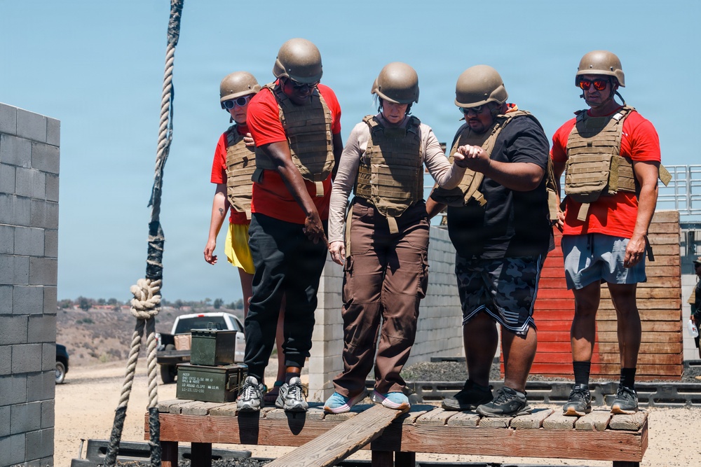 A group of five individuals wearing tactical gear including helmets and plate carriers are participating in what appears to be a military-style obstacle course or training exercise. They are standing on an elevated wooden platform, seemingly navigating a balance or team coordination challenge. The setting appears to be an outdoor training facility in an arid environment.