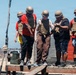 A group of five individuals wearing tactical gear including helmets and plate carriers are participating in what appears to be a military-style obstacle course or training exercise. They are standing on an elevated wooden platform, seemingly navigating a balance or team coordination challenge. The setting appears to be an outdoor training facility in an arid environment.