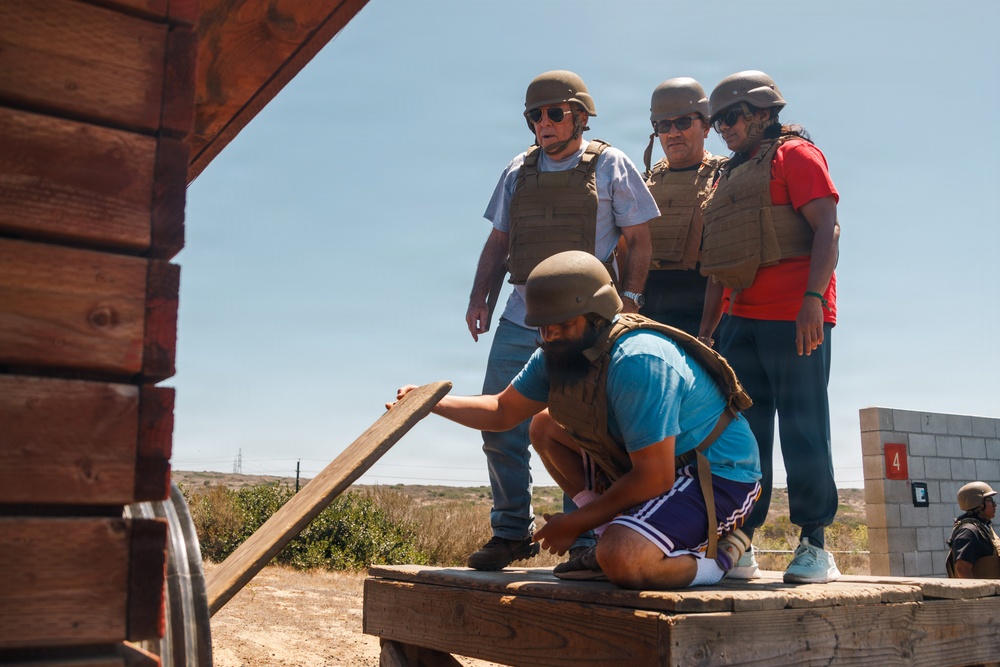 Four individuals wearing tactical helmets and body armor are gathered on a wooden platform in an arid outdoor setting. One person is crouching down and handling a wooden plank, while three others stand and observe. The setting appears to be a training range or tactical exercise area, indicated by numbered concrete block structures visible in the background.