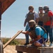 Four individuals wearing tactical helmets and body armor are gathered on a wooden platform in an arid outdoor setting. One person is crouching down and handling a wooden plank, while three others stand and observe. The setting appears to be a training range or tactical exercise area, indicated by numbered concrete block structures visible in the background.