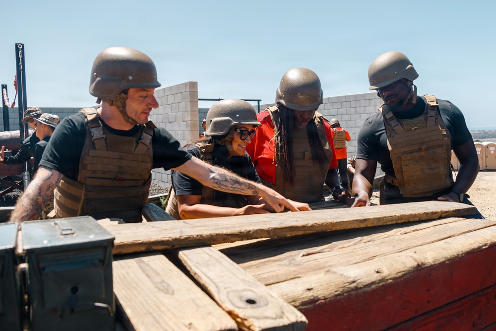 Four individuals wearing military-style tactical gear including helmets and plate carrier vests are gathered around a wooden structure or platform in an outdoor setting. They appear to be engaged in a team activity or challenge, possibly a military-style training exercise or reality competition. The setting includes a concrete block wall, sandy ground, and portable toilets visible in the background.