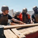 Four individuals wearing military-style tactical gear including helmets and plate carrier vests are gathered around a wooden structure or platform in an outdoor setting. They appear to be engaged in a team activity or challenge, possibly a military-style training exercise or reality competition. The setting includes a concrete block wall, sandy ground, and portable toilets visible in the background.