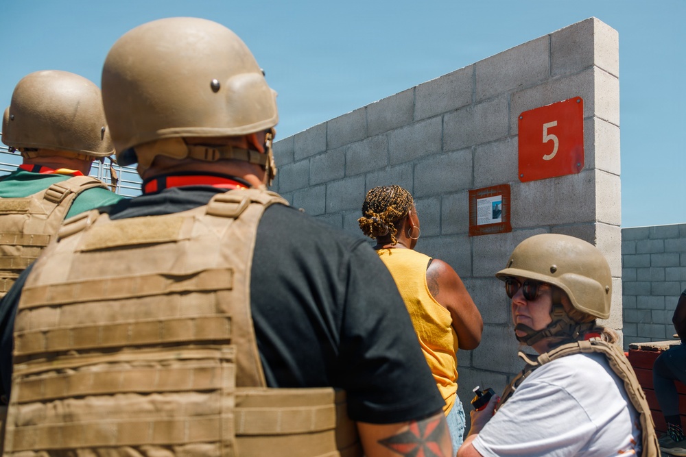 Several individuals are gathered near a concrete block wall, with at least two wearing tactical gear including helmets and plate carrier vests. A red sign with the number '5' is mounted on the wall alongside a smaller informational placard. The setting appears to be an outdoor area under clear blue skies.