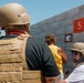 Several individuals are gathered near a concrete block wall, with at least two wearing tactical gear including helmets and plate carrier vests. A red sign with the number '5' is mounted on the wall alongside a smaller informational placard. The setting appears to be an outdoor area under clear blue skies.