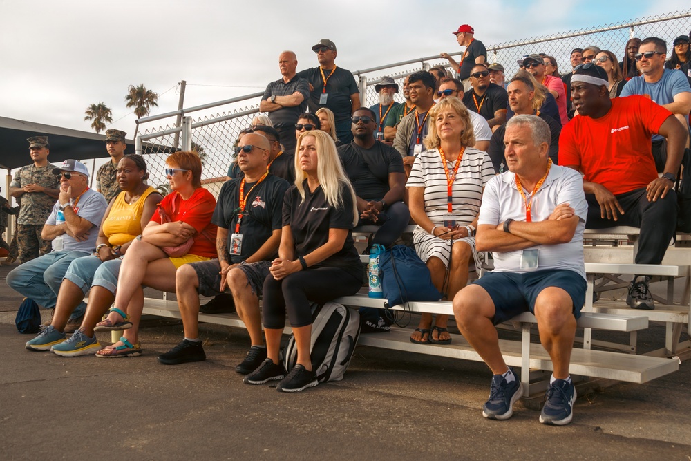 A large group of civilians and military personnel are seated on metal bleachers, apparently watching an event or presentation. Many attendees are wearing orange/red lanyards with credentials, suggesting an organized official visit or demonstration. The crowd appears attentive and focused on something outside the frame.