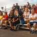 A large group of civilians and military personnel are seated on metal bleachers, apparently watching an event or presentation. Many attendees are wearing orange/red lanyards with credentials, suggesting an organized official visit or demonstration. The crowd appears attentive and focused on something outside the frame.