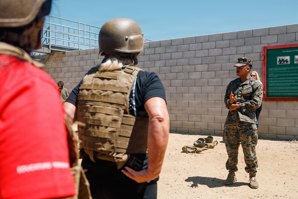 A military briefing or orientation is taking place outdoors at what appears to be a training facility or base. A uniformed Marine in MARPAT camouflage is addressing a small group of civilians, one of whom is wearing a tactical vest and combat helmet. A green informational sign on the wall behind the Marine references 'The Battle of Fa...' (partially visible), suggesting a historical or educational context.