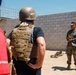 A military briefing or orientation is taking place outdoors at what appears to be a training facility or base. A uniformed Marine in MARPAT camouflage is addressing a small group of civilians, one of whom is wearing a tactical vest and combat helmet. A green informational sign on the wall behind the Marine references 'The Battle of Fa...' (partially visible), suggesting a historical or educational context.