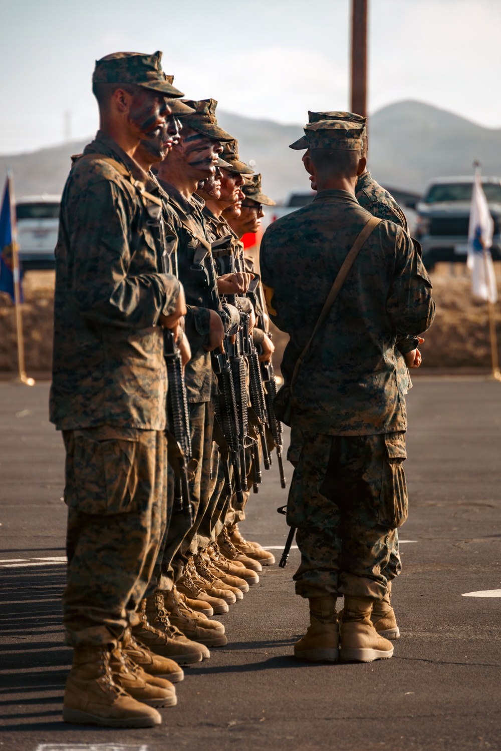 A group of U.S. Marines in MARPAT camouflage uniforms with combat face paint stand in formation while being inspected by a senior Marine. The Marines are armed with M4/M16-style rifles and appear to be at an outdoor military installation with mountains visible in the background. The scene suggests a pre-mission inspection or formal review ceremony.