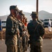 A group of U.S. Marines in MARPAT camouflage uniforms with combat face paint stand in formation while being inspected by a senior Marine. The Marines are armed with M4/M16-style rifles and appear to be at an outdoor military installation with mountains visible in the background. The scene suggests a pre-mission inspection or formal review ceremony.