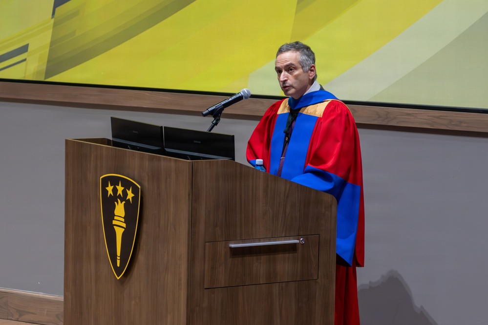 A middle-aged man wearing academic regalia in red, blue, and gold stands at a wooden podium delivering a speech. The podium bears a gold shield emblem featuring a torch and three stars, suggesting a university or academic institution. The setting appears to be a formal academic ceremony such as a commencement or convocation.