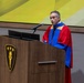 A middle-aged man wearing academic regalia in red, blue, and gold stands at a wooden podium delivering a speech. The podium bears a gold shield emblem featuring a torch and three stars, suggesting a university or academic institution. The setting appears to be a formal academic ceremony such as a commencement or convocation.