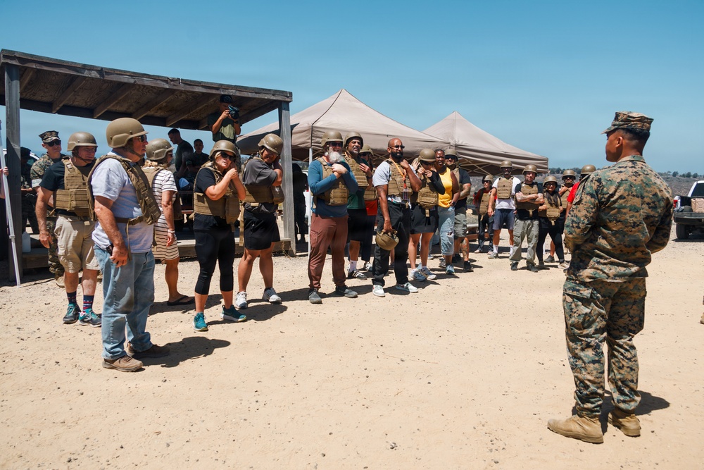 A U.S. Marine in full camouflage uniform addresses a large group of civilians wearing borrowed tactical gear including body armor and helmets. The setting appears to be a military base training area or range, likely during a civilian orientation or community outreach event. The group is diverse in age and appearance, standing in an open sandy area with canopy tents and a covered structure visible in the background.