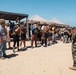 A U.S. Marine in full camouflage uniform addresses a large group of civilians wearing borrowed tactical gear including body armor and helmets. The setting appears to be a military base training area or range, likely during a civilian orientation or community outreach event. The group is diverse in age and appearance, standing in an open sandy area with canopy tents and a covered structure visible in the background.