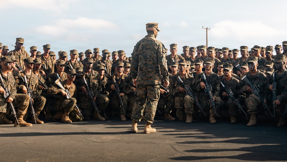 A military leader stands with their back to the camera, addressing a large formation of uniformed service members who are crouched or kneeling on a paved surface. The assembled personnel are wearing woodland MARPAT camouflage uniforms and patrol caps, consistent with U.S. Marine Corps dress, and are armed with rifles. The scene appears to be a pre-mission briefing or unit address conducted outdoors.
