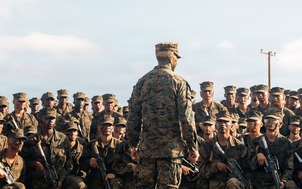 A military leader stands with their back to the camera, addressing a large formation of uniformed service members who are crouched or kneeling on a paved surface. The assembled personnel are wearing woodland MARPAT camouflage uniforms and patrol caps, consistent with U.S. Marine Corps dress, and are armed with rifles. The scene appears to be a pre-mission briefing or unit address conducted outdoors.