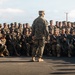 A military leader stands with their back to the camera, addressing a large formation of uniformed service members who are crouched or kneeling on a paved surface. The assembled personnel are wearing woodland MARPAT camouflage uniforms and patrol caps, consistent with U.S. Marine Corps dress, and are armed with rifles. The scene appears to be a pre-mission briefing or unit address conducted outdoors.