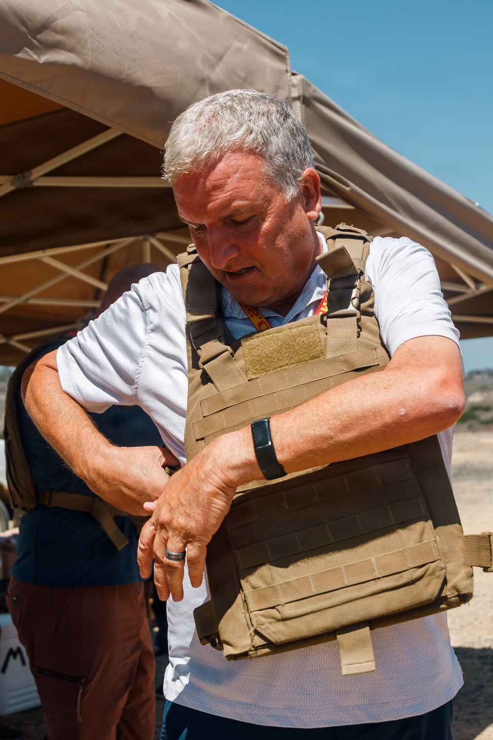 A middle-aged man with gray hair is putting on or adjusting a tan/coyote-colored tactical plate carrier vest over a white polo shirt. He appears to be at an outdoor event or demonstration, standing under a tan canopy tent in what looks like a dry, arid environment. He is looking down at the vest while adjusting it with both hands.