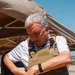 A middle-aged man with gray hair is putting on or adjusting a tan/coyote-colored tactical plate carrier vest over a white polo shirt. He appears to be at an outdoor event or demonstration, standing under a tan canopy tent in what looks like a dry, arid environment. He is looking down at the vest while adjusting it with both hands.
