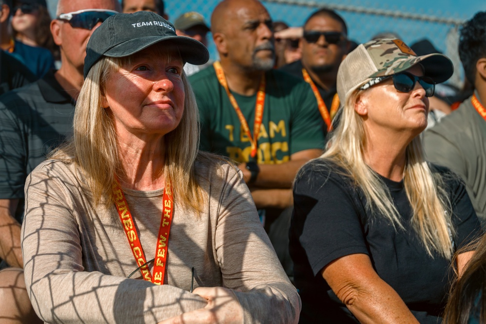 A crowd of people attending what appears to be a Team Rubicon event, identifiable by the organization's branding on hats, lanyards, and shirts. Two women in the foreground are prominently featured, both wearing baseball caps and lanyards, looking attentively in the same direction. The gathering appears to be an outdoor event in bright sunlight.