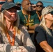 A crowd of people attending what appears to be a Team Rubicon event, identifiable by the organization's branding on hats, lanyards, and shirts. Two women in the foreground are prominently featured, both wearing baseball caps and lanyards, looking attentively in the same direction. The gathering appears to be an outdoor event in bright sunlight.