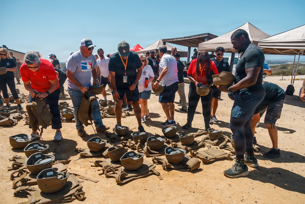 A group of civilians are gathered outdoors on a dirt surface, picking up and examining military-style helmets and gear spread across the ground. The setting appears to be a military base or training facility, likely during a civilian orientation or community outreach event. Event tents and a coastal landscape are visible in the background.