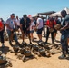 A group of civilians are gathered outdoors on a dirt surface, picking up and examining military-style helmets and gear spread across the ground. The setting appears to be a military base or training facility, likely during a civilian orientation or community outreach event. Event tents and a coastal landscape are visible in the background.