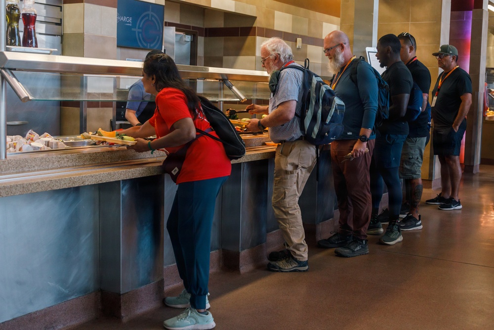 A group of people are standing in line at a cafeteria or food service counter, selecting items from a buffet-style serving station. A staff member in blue is visible behind the counter serving food. The setting appears to be a modern institutional dining facility, possibly at a convention center, stadium, or large venue.