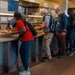 A group of people are standing in line at a cafeteria or food service counter, selecting items from a buffet-style serving station. A staff member in blue is visible behind the counter serving food. The setting appears to be a modern institutional dining facility, possibly at a convention center, stadium, or large venue.