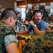 A group of military personnel in camouflage uniforms are eating together in what appears to be a military dining facility (mess hall). One civilian man in a gray t-shirt sits among the uniformed service members, engaging in conversation. The setting suggests a communal meal in a structured military environment.
