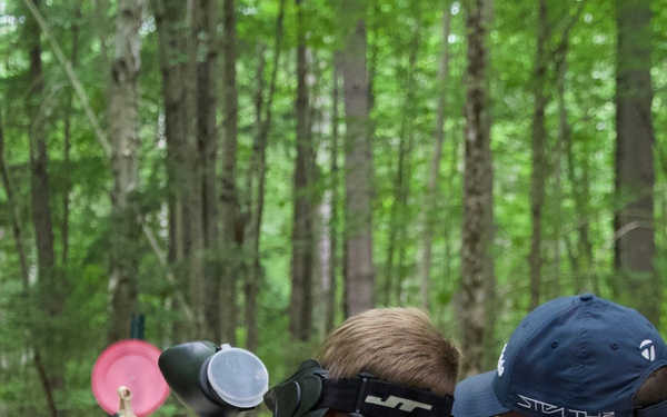 A young child wearing a JT brand protective mask and purple sweatshirt is aiming a paintball marker at a pink disc target in a wooded outdoor setting. An adult male wearing a blue 'Stealth' cap and red shirt is seated nearby, supervising the activity. The scene appears to be a supervised paintball shooting range set up in a forested area.
