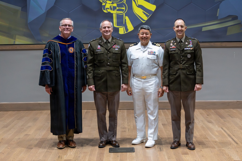 Four individuals pose for a formal group photograph in what appears to be an academic or ceremonial venue. The group includes one person in academic doctoral regalia and three individuals in military dress uniforms representing different branches of service. The setting features a decorative blue and gold mural backdrop and a hardwood floor stage.