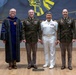 Four individuals pose for a formal group photograph in what appears to be an academic or ceremonial venue. The group includes one person in academic doctoral regalia and three individuals in military dress uniforms representing different branches of service. The setting features a decorative blue and gold mural backdrop and a hardwood floor stage.