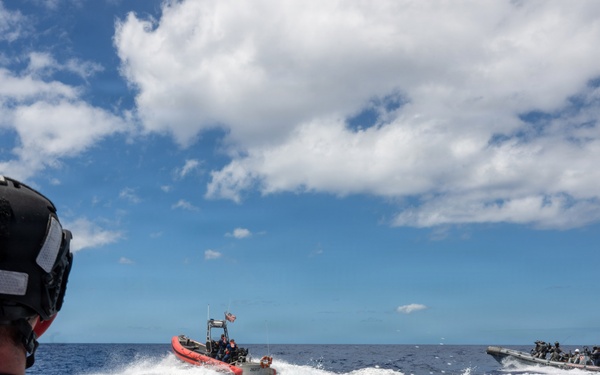 A U.S. Coast Guard member wearing an orange vest and black helmet observes two rigid inflatable boats (RIBs) speeding across open ocean water. One boat is orange and one is gray/camouflage, both carrying multiple personnel and creating large wakes. The scene appears to depict a maritime interdiction or training operation in open blue water under partly cloudy skies.