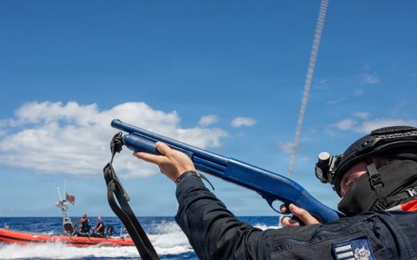 A uniformed maritime law enforcement officer is aiming a blue-colored shotgun (likely a training/less-lethal weapon) from a vessel during what appears to be a maritime interdiction or training exercise at sea. A red inflatable rigid-hull inflatable boat (RHIB) with personnel aboard is visible in the background. The officer is wearing tactical gear including a helmet, face covering, body armor, and an orange life vest.