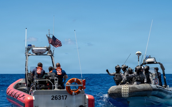 Two rigid-hull inflatable boats (RHIBs) are operating in open ocean waters. The left vessel, numbered 26317, flies a U.S. flag and carries personnel in orange life vests consistent with U.S. Coast Guard markings. The right vessel carries multiple personnel in dark tactical gear and helmets, suggesting a joint maritime law enforcement or training operation.
