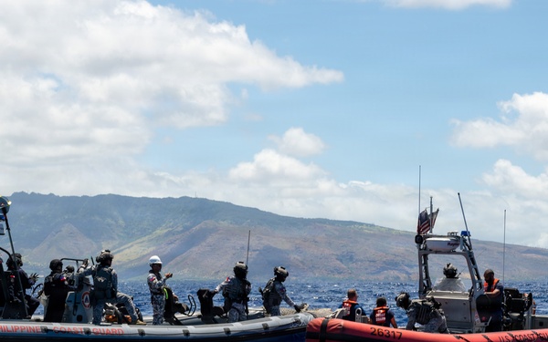 Two rigid inflatable boats (RIBs) are operating side by side in open blue ocean waters with a mountainous coastline visible in the background. The left vessel is marked 'Philippine Coast Guard' and the right vessel is marked 'U.S. Coast Guard' with hull number 26317 and flies an American flag. Personnel in tactical gear are visible aboard both vessels, suggesting a joint maritime training exercise or cooperative patrol operation.