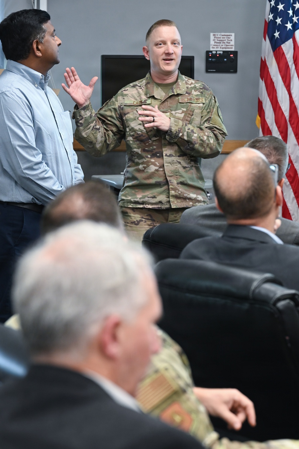 A U.S. Air Force servicemember in OCP (Operational Camouflage Pattern) uniform is speaking or presenting to a seated audience in what appears to be a briefing room or conference facility. He is standing alongside a civilian in a light blue shirt, both facing the audience. An American flag is visible in the background.