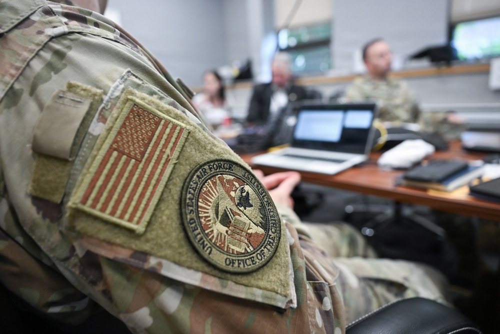 A close-up photograph focuses on the shoulder of a U.S. Air Force personnel wearing OCP (Operational Camouflage Pattern) uniform, displaying a subdued American flag patch and a 'United States Air Force - Office of the Chief' circular patch. In the blurred background, several individuals including military and civilian personnel are seated around a conference table with laptops open, suggesting a briefing or meeting. The image emphasizes the insignia while providing context of an official military office environment.