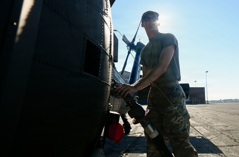 A military service member in camouflage uniform is performing maintenance or refueling operations on a dark-colored military helicopter on a tarmac. The image is backlit by bright sunlight, creating a dramatic silhouette effect. The setting appears to be a military airfield or base with hangar structures visible in the background.