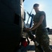 A military service member in camouflage uniform is performing maintenance or refueling operations on a dark-colored military helicopter on a tarmac. The image is backlit by bright sunlight, creating a dramatic silhouette effect. The setting appears to be a military airfield or base with hangar structures visible in the background.